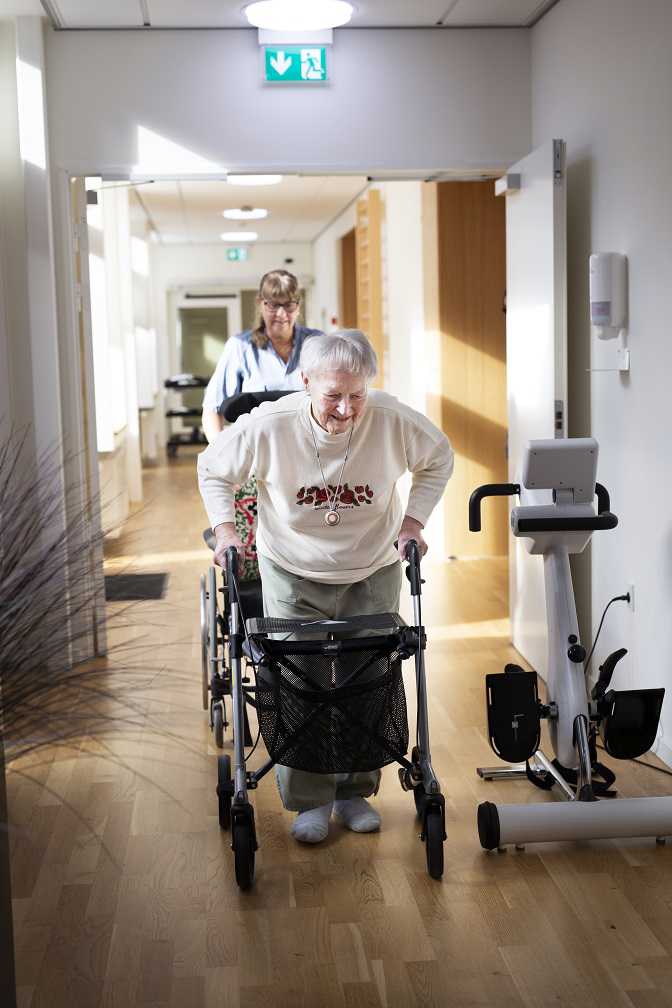 Ældre dame går ned ad en gang. Der er en SOSU-assistent, som følger efter og hjælper hende.