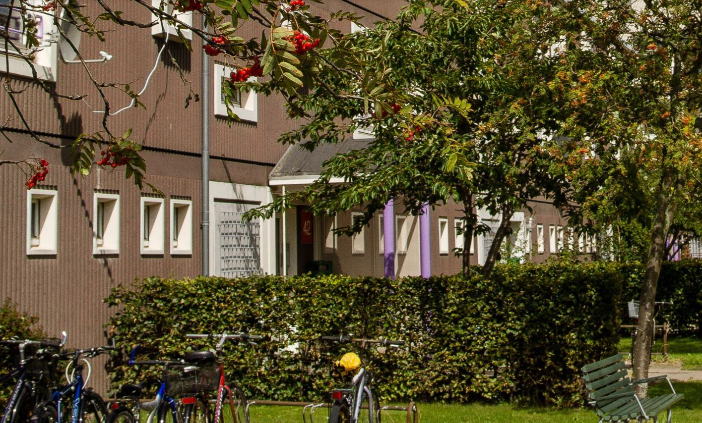 Integrationsboliger "Roen". Det er en brunlig bygning med masser af grøn natur foran. 